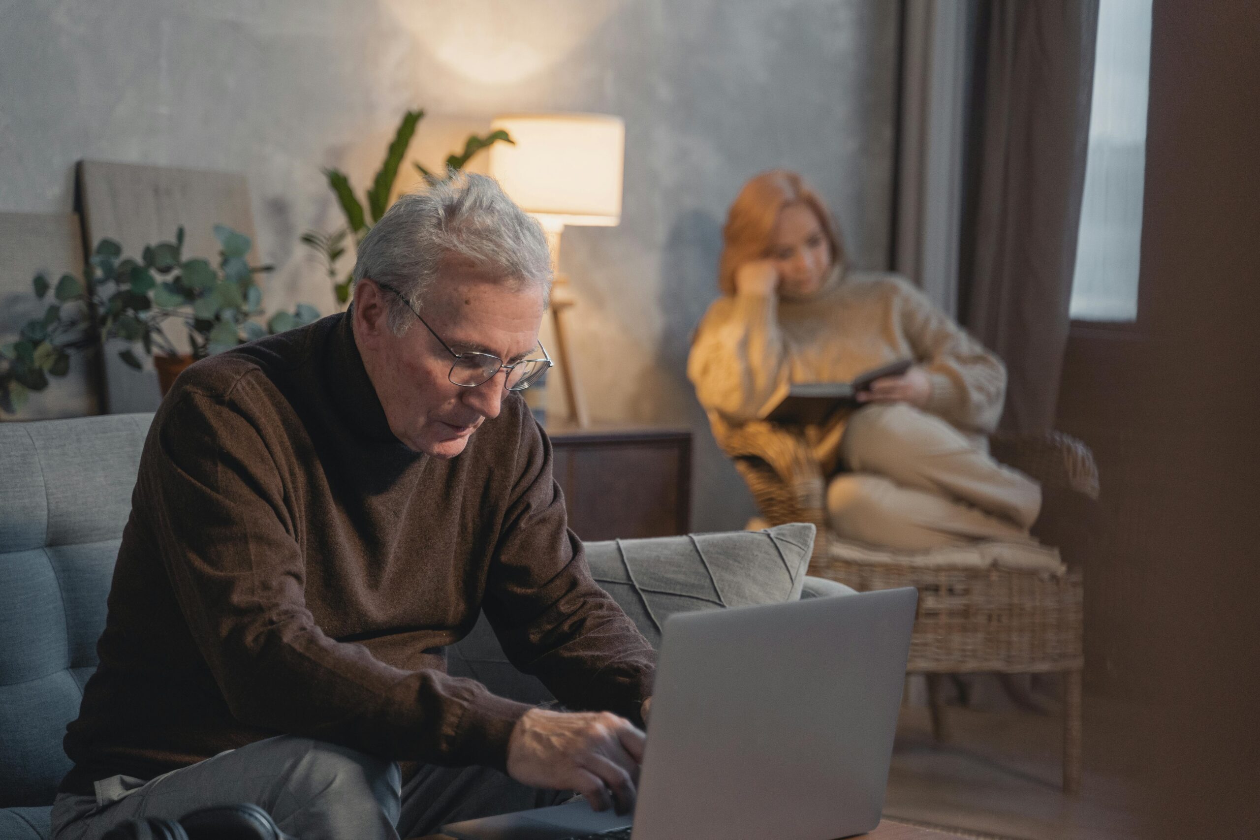 Elderly man using a laptop at home while a woman reads in the background